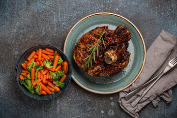 Delicious roasted ham or pork knuckle with glazed brown crispy skin, red pepper berries and rosemary on plate, steamed vegetables on rustic stone background top view, traditional and festive meal