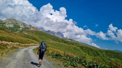 Fototapeta premium A man hiking along a gravelled road in high Caucasus mountains in Georgia. The road leads to a ski resort on a steep slope. Thick clouds in the back. Lush pastures on the sides. Barren peaks.