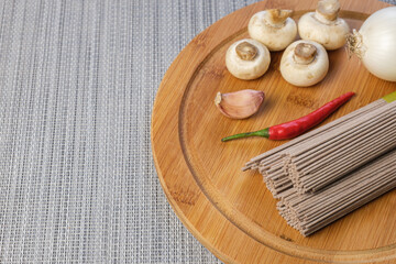 Dry Soba buckwheat noodles and ingredients for preparing an Asian dish. Soba buckwheat noodles, champignons, hot pepper pods, garlic, onion on cutting board. Top view, copy space