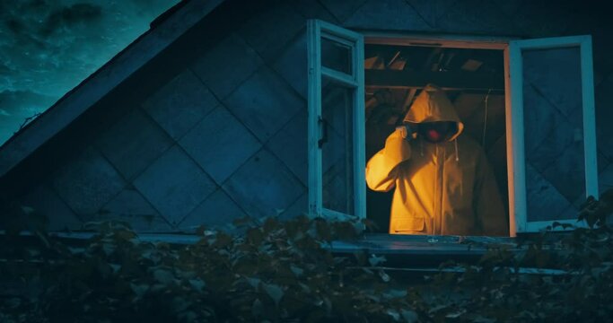 Maniac in a yellow raincoat with an axe and a knife on the top floor of an old house. The concept of horror, thriller and Halloween holiday. At night, the camera shoots outside through the window