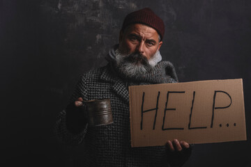 Portrait of poor homeless man with poster and a pot for alms on a dark background