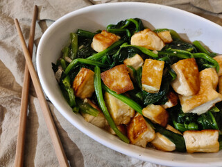Dish made of fried tofu with boiled pak choi served in oval bowl. There is a pair of chopsticks next to the bowl and everything sits on brown table cloth.