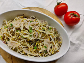 Cold fresh salad made of mungo sprouts, corn, parsley, and onion served in white oval bowl. There are two whole tomatoes next to the bowl.