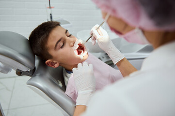 Confident child boy sitting in dentist's chair in contemporary dental clinic while unrecognizable female dentist doing medical manipulations in his opened mouth by cheek retractor and dental mirror