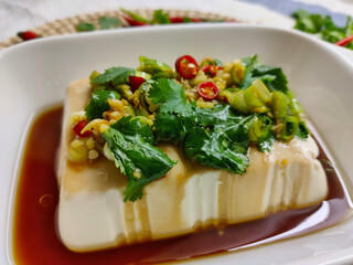 Silken tofu prepared in soy sauce with coriander and chili pepper on top of it. It's served in white rectangle bowl. There is also decoration around the bowl in blured area.