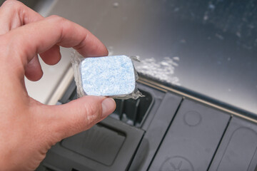 A hand putting blue dishwasher soap tablet in a water-soluble packaging in a dishashind machine...
