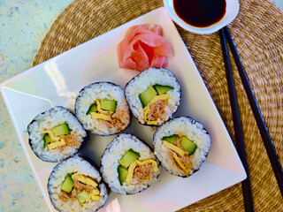 Nicely decorated plate with six pieces of sushi with cucumber, tuna, and egg in it. There is also few pieces of ping pickled ginger. Next to the plate is a bowl with soy sauce and pair of chopsticks.