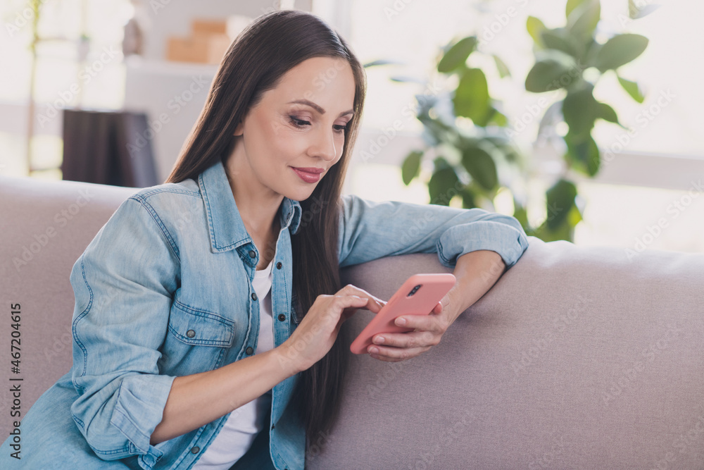 Sticker Portrait of attractive focused cheery woman influencer sitting on divan using device post smm at home indoors