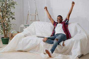 Joyful dark-skinned student sits on couch secured with plastic sheeting to prevent him from getting dirty during renovation of apartment raises hands in air with happiness victory achievement of goal