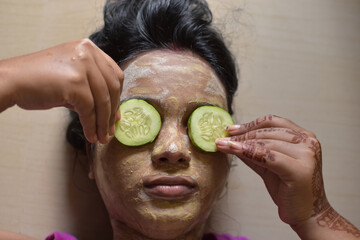 applying facial pack mask gel on face of a lady eyes closed with cucumber pieces.