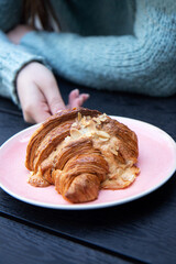 Delicious and tasty croissants with slices nuts in pink plate on black wood background and female hand in a cozy blue sweater