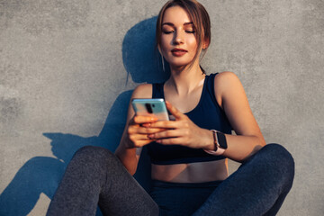 Sports fitness woman using her mobile phone during a break from workout while sitting on a gray...