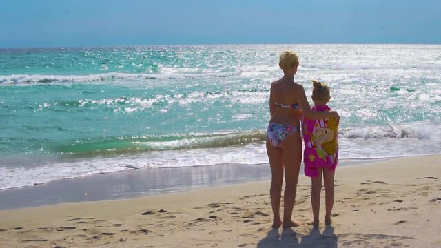 Mom and daughter at sea look naa avolna stand with their backs and look at the beautiful tide, the wind blows, in the heat of summer in swimsuits