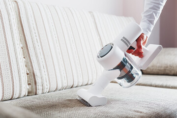 A man's hand holds a modern wireless vacuum cleaner for cleaning the sofa in the house. - obrazy, fototapety, plakaty
