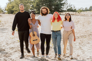 A group of very good friends are standing on the beach by the lake, of different nationalities, skin colors, holding beer bottles in their hands, playing guitar, having fun in each other's company