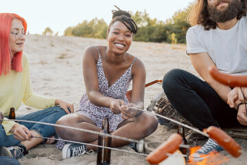 Beautiful black woman with white smile dreadlocks tied up in bun is sitting with friends at bonfire on sand frying sausages bread on sticks meeting in open air, drinking beer from bottles