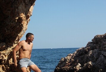 Topless man in blue shorts sitting on rock formation near sea