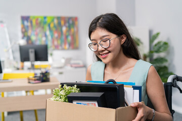 Positive smiling cheerful attractive young girl with glasses is disabled rides in wheelchair and because of this gets promotion to another position, holds on lap cardboard box with things accessories