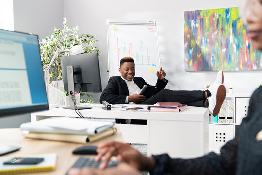 The dark-skinned handsome boss of company sits comfortably in chair with feet on desk leans on it quickly comes up with a brilliant idea rises up holding the tablet in hand gestures