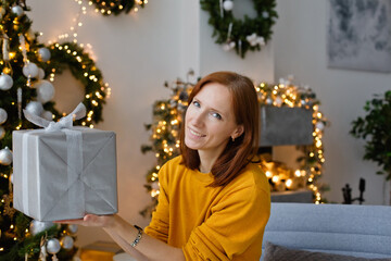 A young woman in a yellow sweater is sitting with a gift in a beautifully decorated room for Christmas. Hiding behind a gift, holding it in front of him.