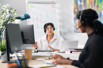 Female co-workers are on break sitting at desks with headphones on their ears in front of a computer waiting for calls to come in, gossiping among themselves, call center, insurance company