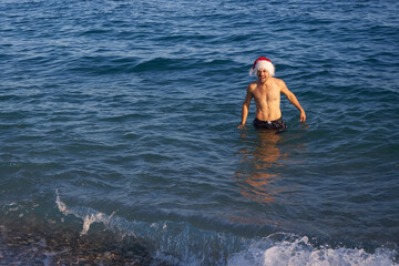 Young man in Christmas hat staying near the beach. Tropical sandy beach seasonal travel vacation, blue sky background outdoors. Back side view of winter holidays vacation