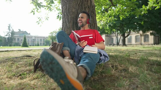Front view happy bearded relaxed student sitting at tree on college yard singing listening to music in earphones. Handsome young Middle Eastern man enjoying hobby studying outdoors in sunshine