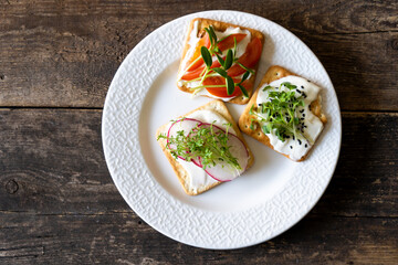 Fototapeta na wymiar Set of Bruschett with cream cheese and tomato spread. Radish, micro green and flaxseed in food. Simple food on the plate. Breakfast in a white plate top view. Wooden background. Cracker
