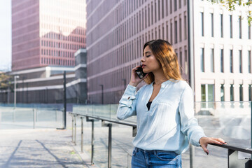 Fototapeta premium Attractive young businesswoman talking on smartphone with business partners outdoors, professional female architect working on new project surrounded by buildings