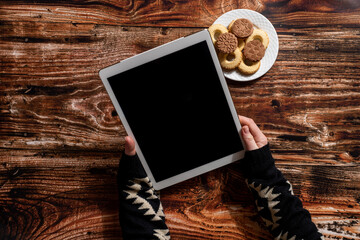 Woman's hands holding digital tablet on the wooden table with a plate full of cookies
