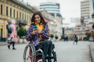 Woman in wheelchair using a smartphone while out in the city - obrazy, fototapety, plakaty