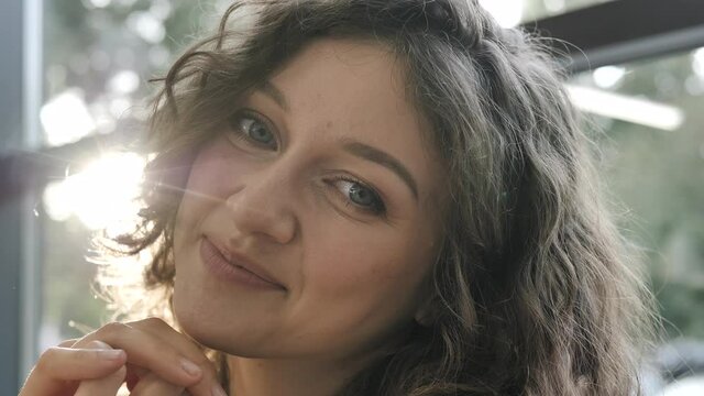 Happy curly haired young woman with light makeup smiles cute and poses looking into camera sitting against window head shot