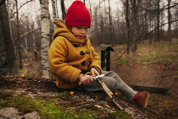 Little Girl Sitting in the Forest and Planing a Stick With a Knife