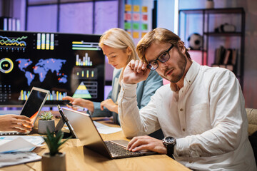 Tensive caucasian man and woman working on financial report during evening time at office. Two partners using modern laptop and tablet. Overwork and deadlines concept.