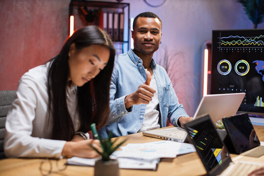 Technology and people concept. African american man and asian woman using laptop and tablet late at office. Positive African man showing thumb up at camera