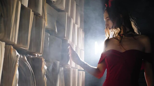 Young woman walks along storage wall with books attached and runs hand over old torn pages slowly at back light