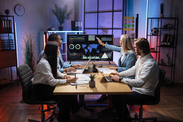 Caucasian woman pointing on monitor with financial report during business meeting with her multi ethnic colleagues. Four partners sitting at desk and discussing common project.