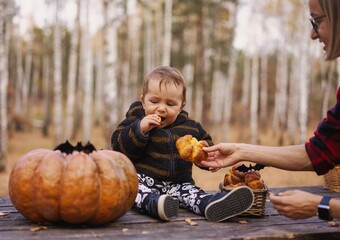 Young woman and her baby son in autumn park, boy playing with helloween pumpkin and eating pumpkin bun