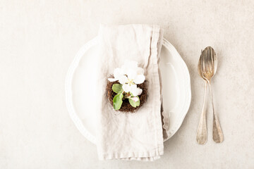 Elegant easter spring table setting design with white blooming flowers, cutlery, empty plate, napkin on beige table. Top view, flat lay