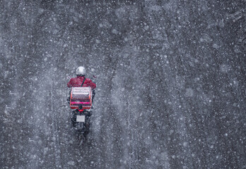 Delivery service from cafes and restaurants, a courier on a scooter with a yellow backpack travels under the snow. A courier delivers food on a motorcycle.