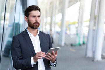 stylish bearded businessman in formal business suit standing working with tablet in hands on background modern office building outside. Man using smartphone or uses mobile phone outdoors city street