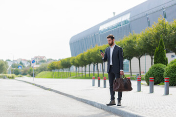 stylish bearded business man with travel bag and mobile phone in hands walks on city street modern urban background. Businessman in a suit goes and uses a smartphone. tourist trip. caucasian manager