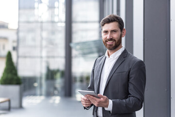 Cheerful and successful businessman works outdoors near the office holds a tablet, smiles reads good news