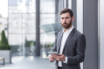 stylish bearded businessman in formal business suit standing working with tablet in hands on background modern office building outside.