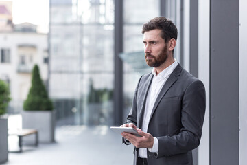 Obraz na płótnie Canvas stylish bearded businessman in formal business suit standing working with tablet in hands on background modern office building outside.