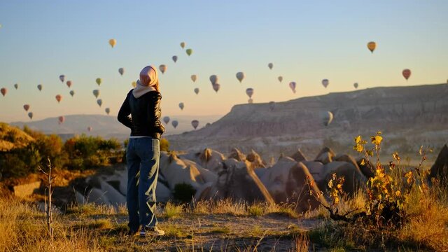 slender attractive young woman at dawn in pink hat, leather jacket and tight bell-bottomed jeans looks at parade of balloons in sky over Turkey. Many balloons with balloonists are navigating in sky.