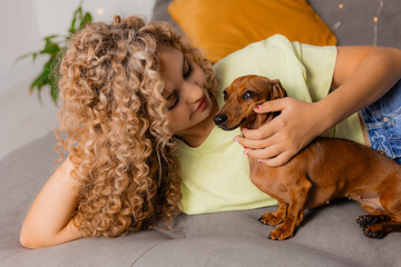 charming blonde girl with curls is lying on a gray bed with a dachshund dog. Happy time with pets. High quality photo