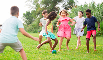 Happy preteen girls and boys of different nationalities playing football on green lawn in spring city park..