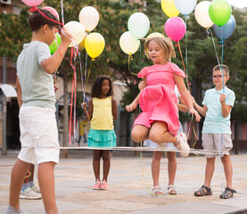 Happy preteen kids of different nationalities holding colorful balloons in hands, skipping on chinese jump rope in courtyard on summer day