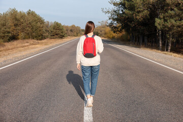 Young woman with backpack going along road near forest, back view